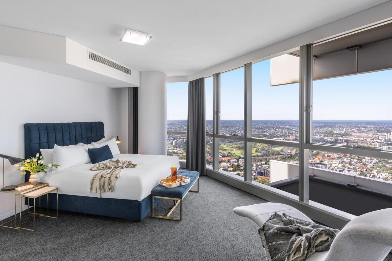 赫歇尔街美利通套房酒店 布里斯班 外观 照片 The photo shows a modern hotel room featuring a large bed with a blue upholstered headboard and white bedding. The room has a stylish design with a gray carpet and large windows that provide a panoramic view of the surrounding landscape. There is a s