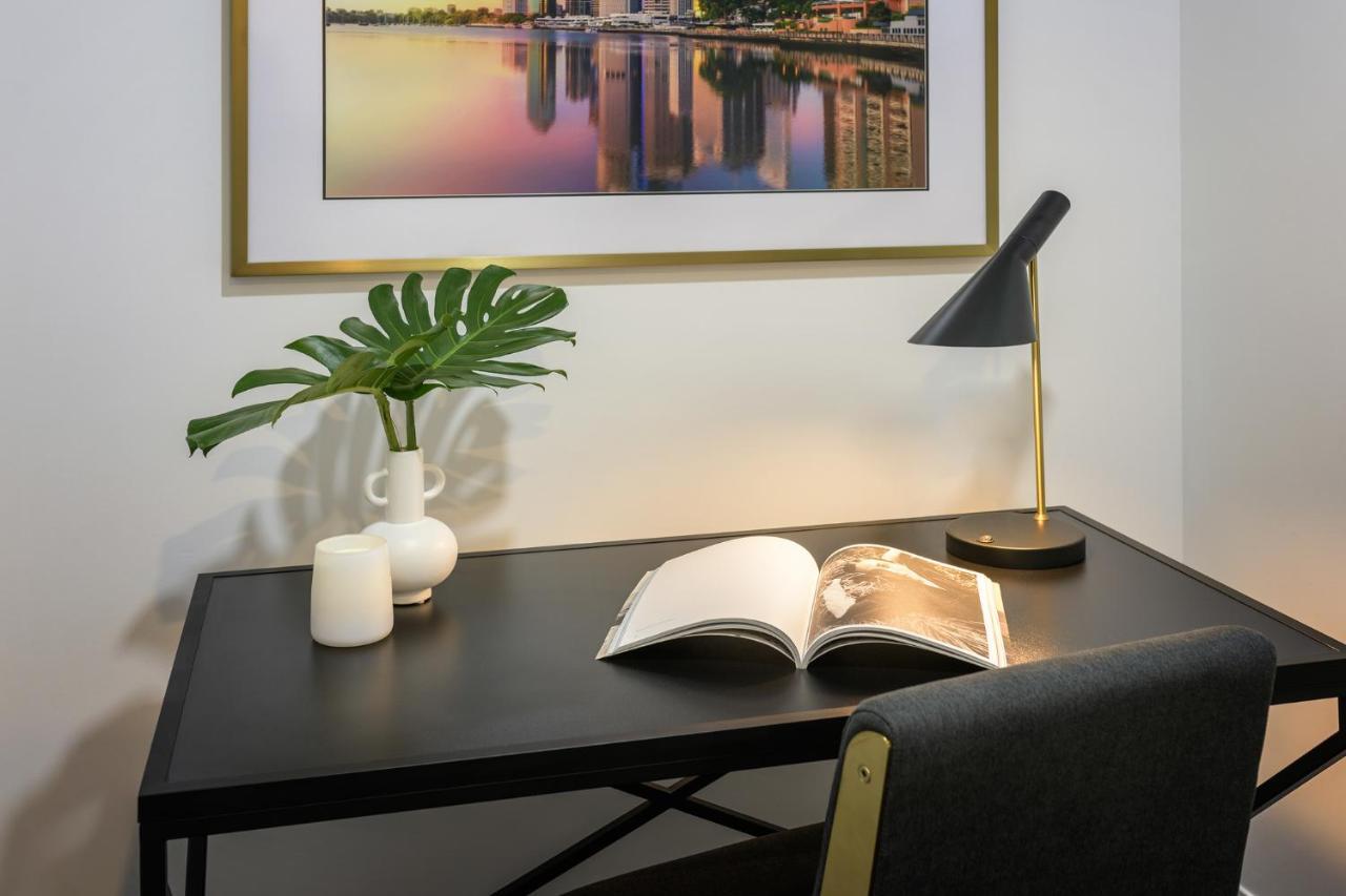 赫歇尔街美利通套房酒店 布里斯班 外观 照片 The image shows a modern workspace featuring a black desk with a small open book on it. A stylish desk lamp provides lighting, and there is a minimalist white vase with a green plant beside it. There are also two small candles on the desk. Above the 