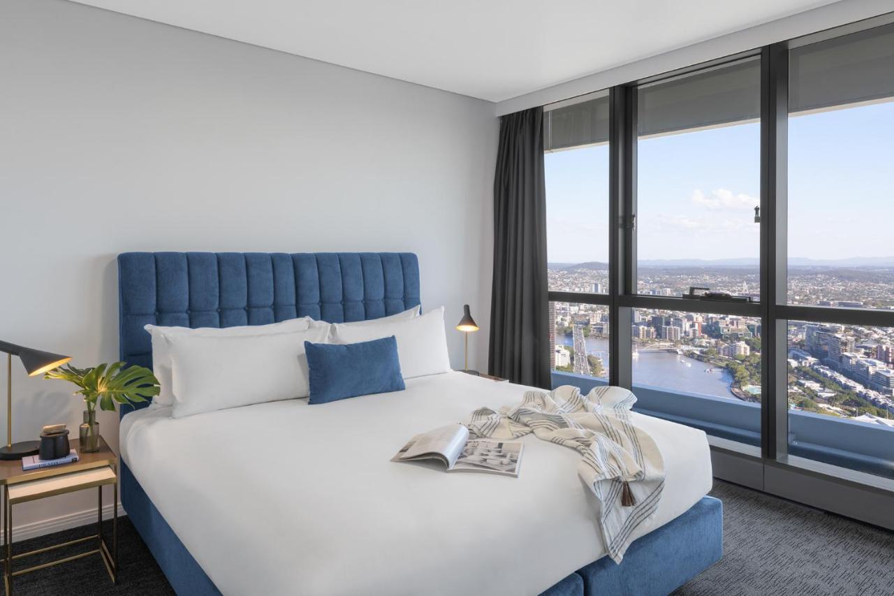 赫歇尔街美利通套房酒店 布里斯班 外观 照片 The photo shows a modern hotel room featuring a neatly made bed with a blue headboard and white bedding. A decorative throw blanket is casually placed on the bed, and there is a small plant on a bedside table. In the background, large windows offer a