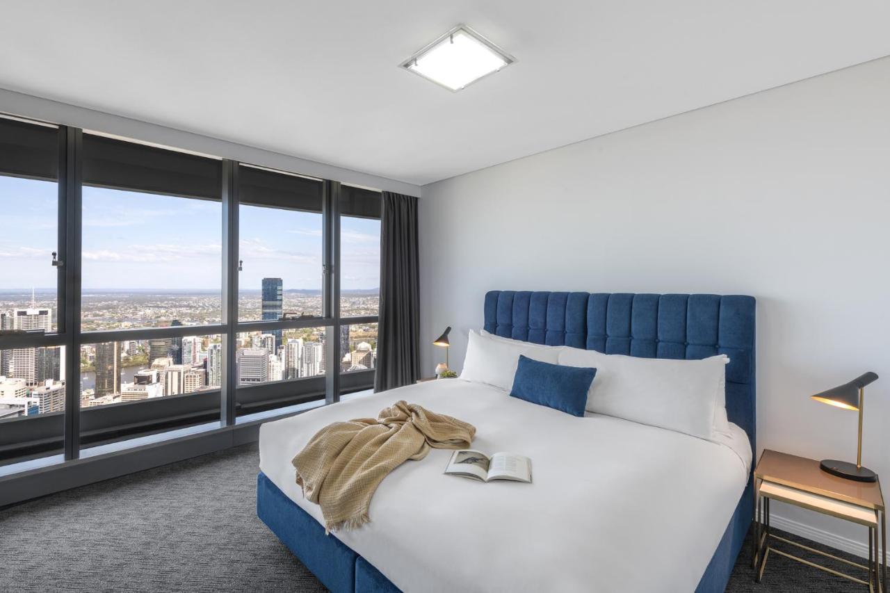 赫歇尔街美利通套房酒店 布里斯班 外观 照片 The image shows a modern hotel bedroom with large windows that offer a view of a cityscape. The room features a spacious bed with a blue upholstered headboard and white bedding, complemented by a beige blanket draped across the bed. On the bed, there
