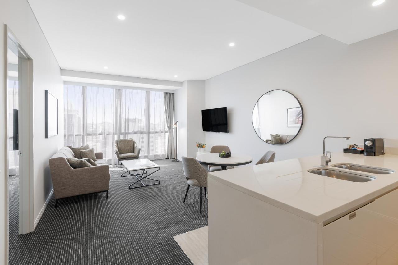 赫歇尔街美利通套房酒店 布里斯班 外观 照片 The photo shows a modern, well-lit living space that appears to be a hotel room or an apartment. It features a cozy seating area with two armchairs and a small coffee table, along with a larger dining table with chairs. There is a round mirror on one