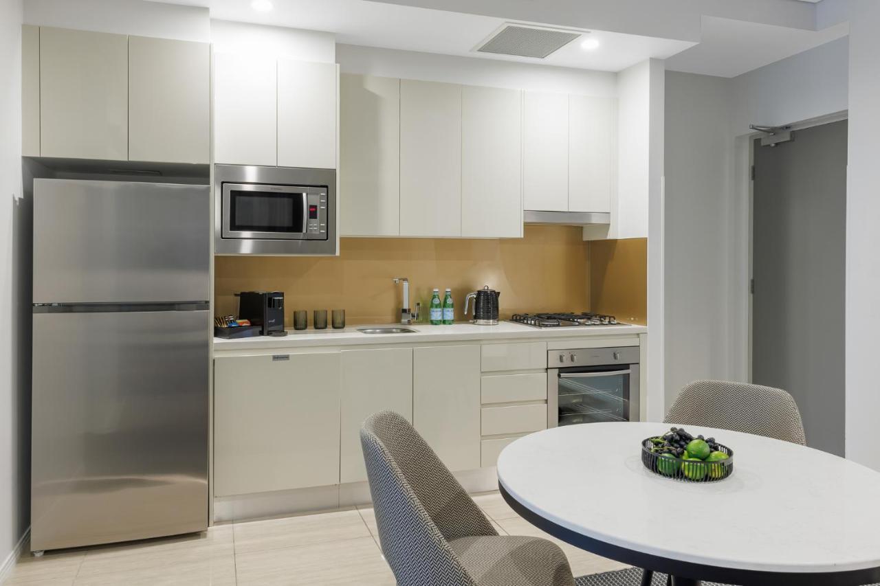 赫歇尔街美利通套房酒店 布里斯班 外观 照片 The photo shows a modern kitchen space featuring sleek white cabinetry and appliances. There is a stainless steel refrigerator and a built-in microwave above a stove. The countertops are light-colored, complementing the overall clean design. A small 