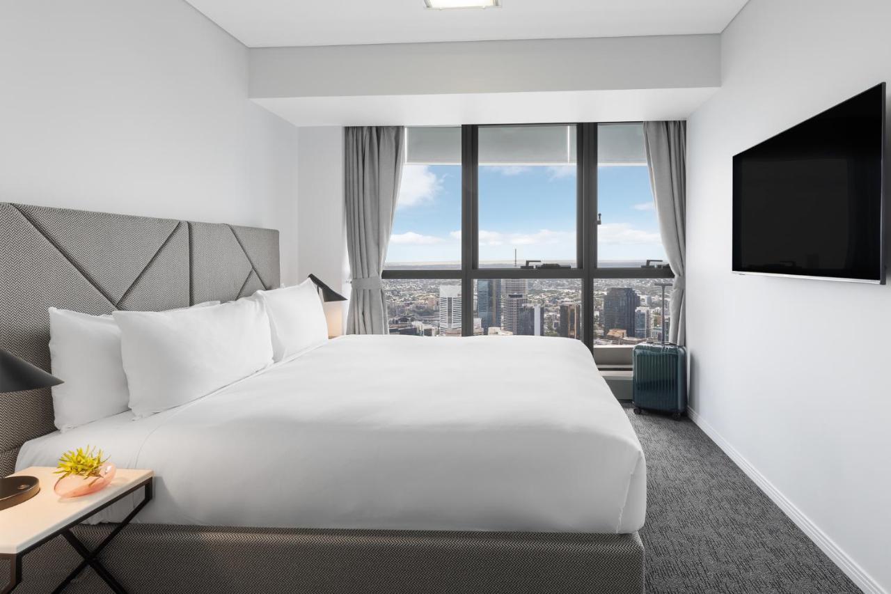 赫歇尔街美利通套房酒店 布里斯班 外观 照片 The photo shows a modern hotel room with a large bed situated against a wall. The bed has white linens and is neatly made. There are two pillows and a decorative throw on the bed. To the side, there is a small nightstand with a decorative item. The r