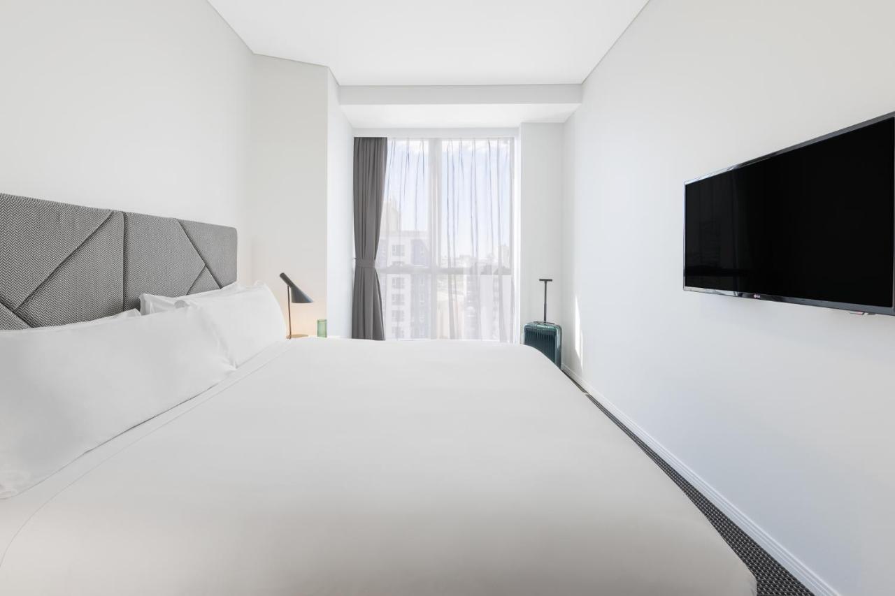 赫歇尔街美利通套房酒店 布里斯班 外观 照片 The photo shows a modern, minimalist hotel room. It features a large bed with a white duvet and pillows against a light-colored wall. To the side, there is a small bedside lamp and a suitcase on a stand. A large window with sheer curtains lets in nat